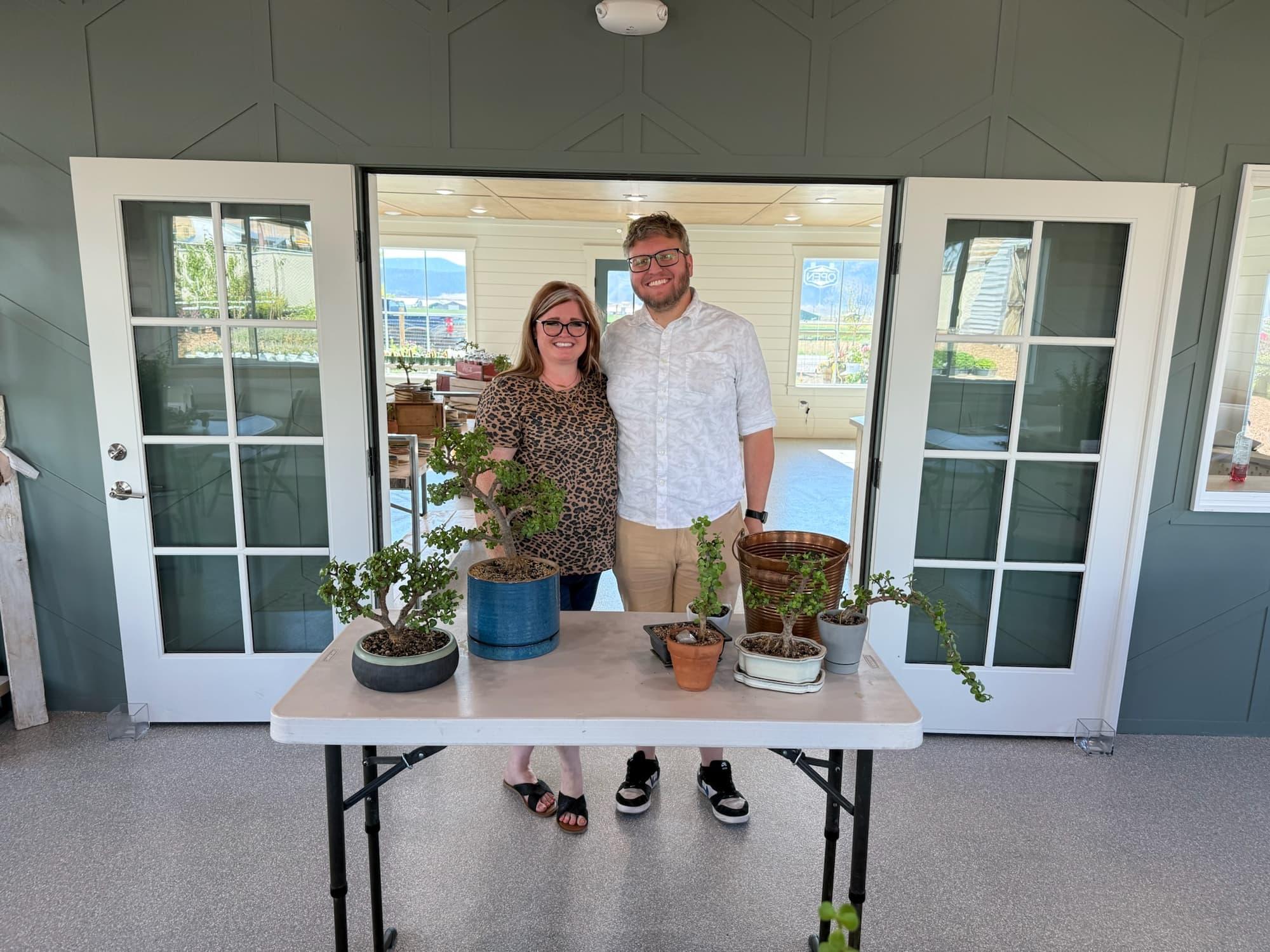 Cass and Landon stand behind a table of dwarf jade bonsai, with Cheesecake in his blue pot, at open French doors