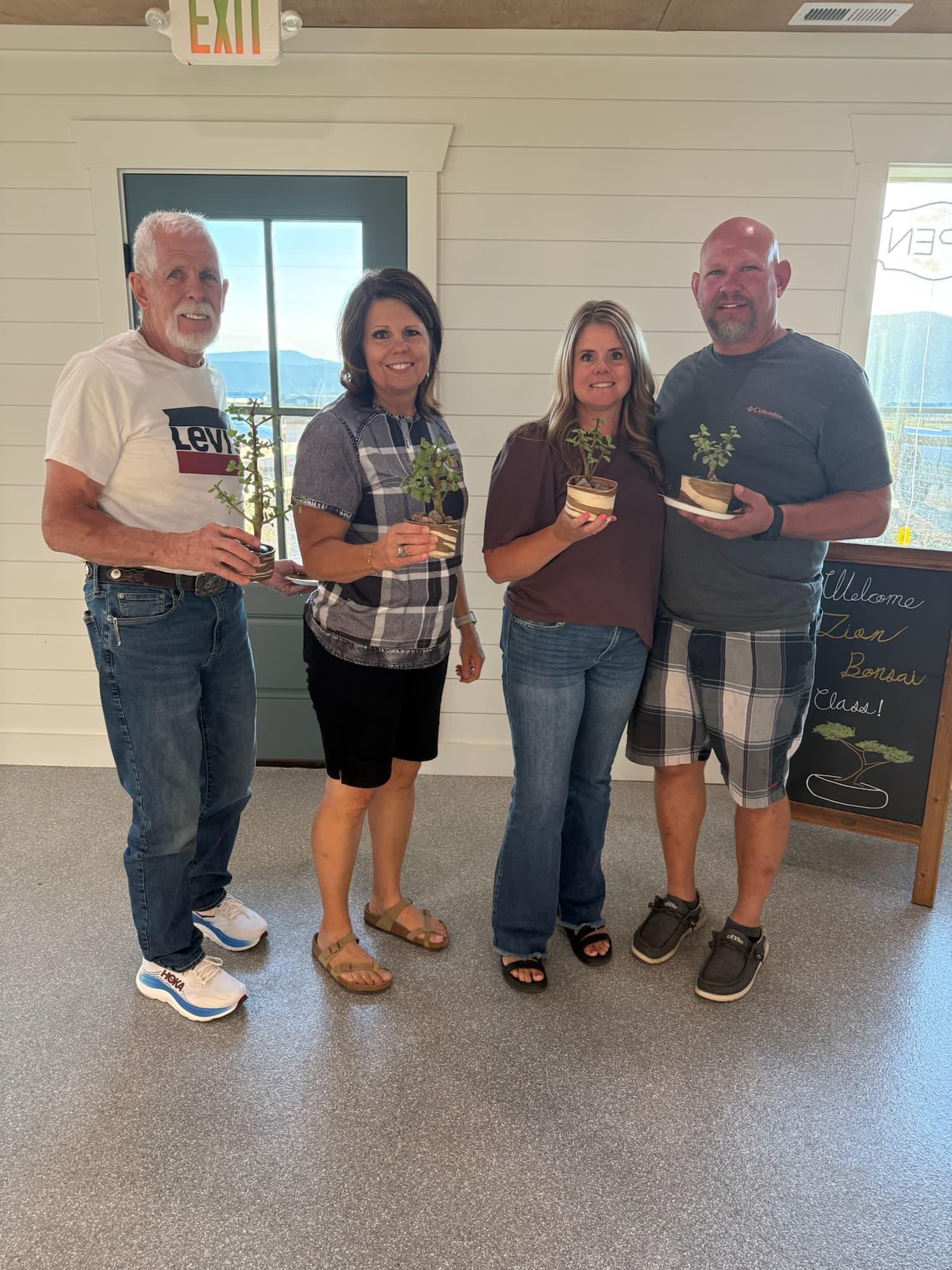 Four participants hold their new dwarf jade bonsai in a white room with a chalkboard sign