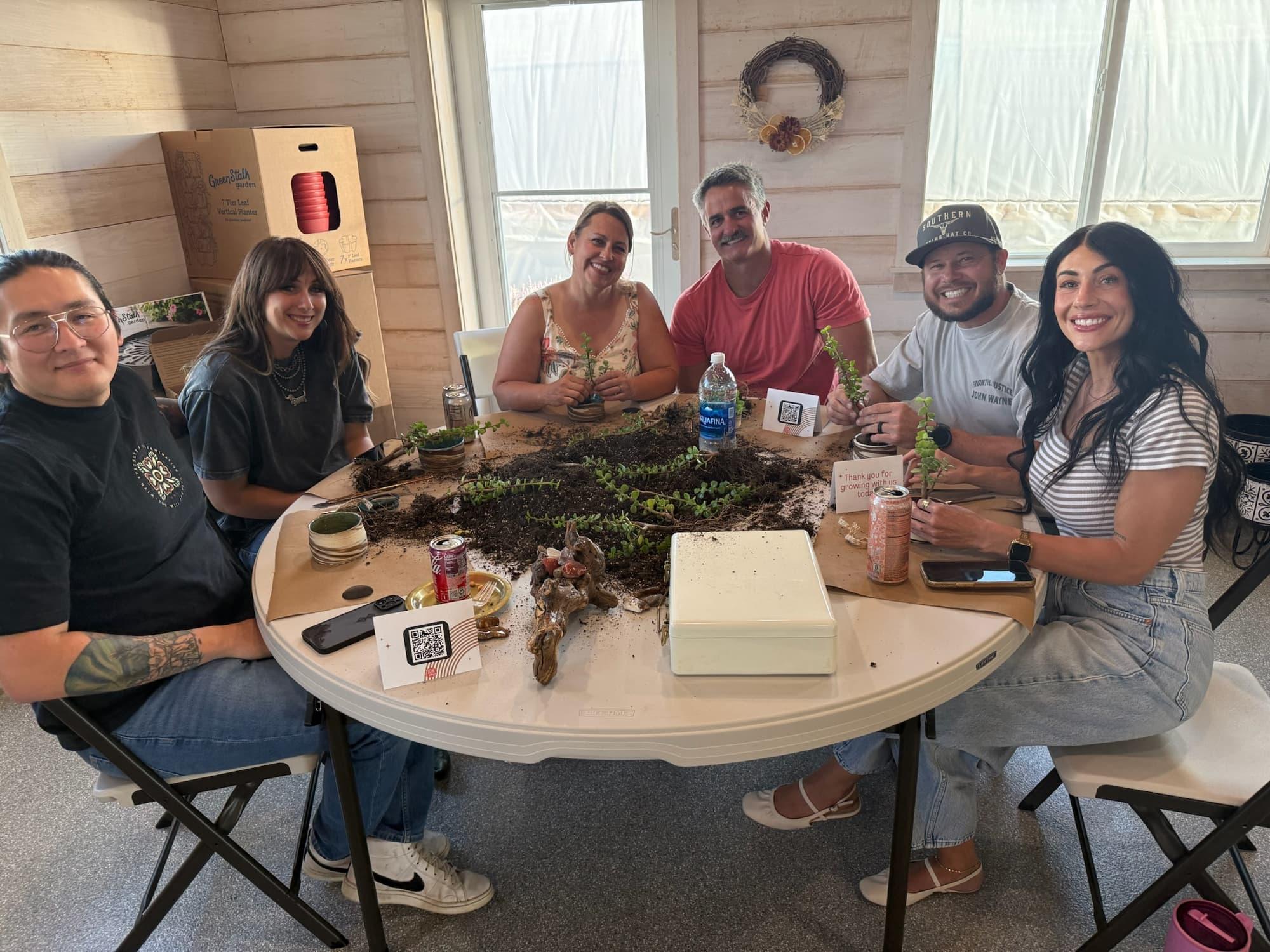 Six participants smile around a round table scattered with soil, dwarf jade cuttings and driftwood