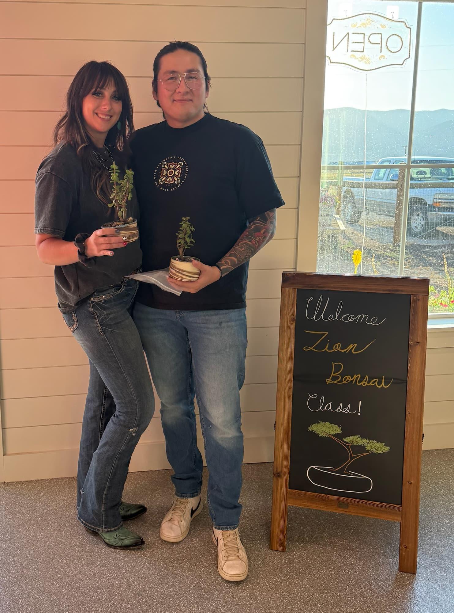 Two participants with small potted dwarf jade bonsai stand by the Zion Bonsai Class chalkboard