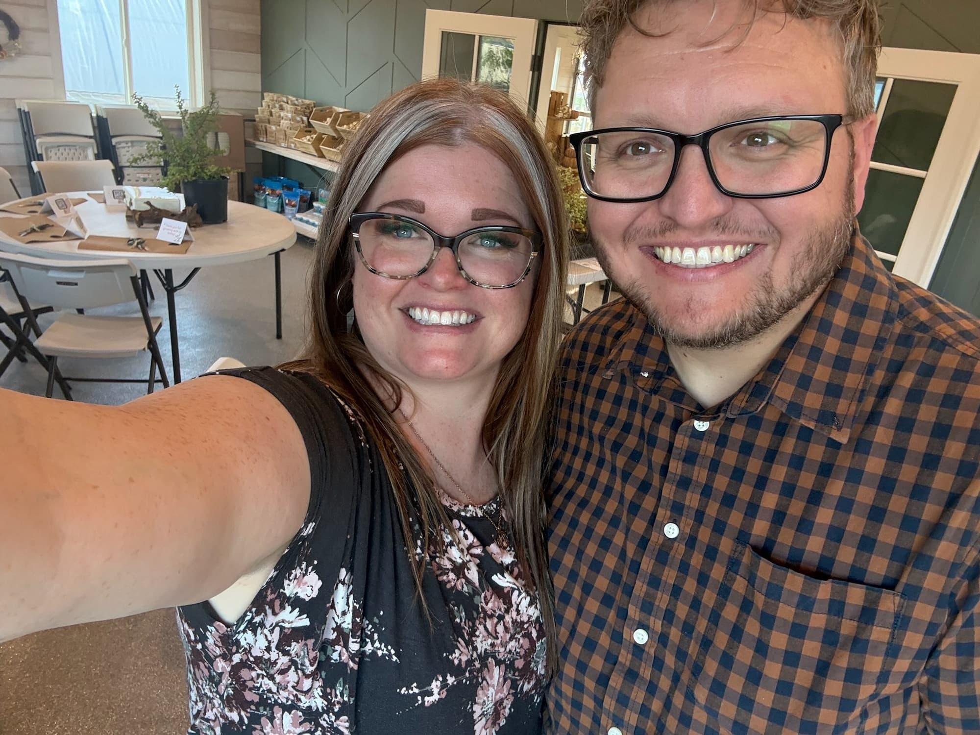 Cass and Landon smile for a selfie in a bright workshop venue, tables set for class behind them